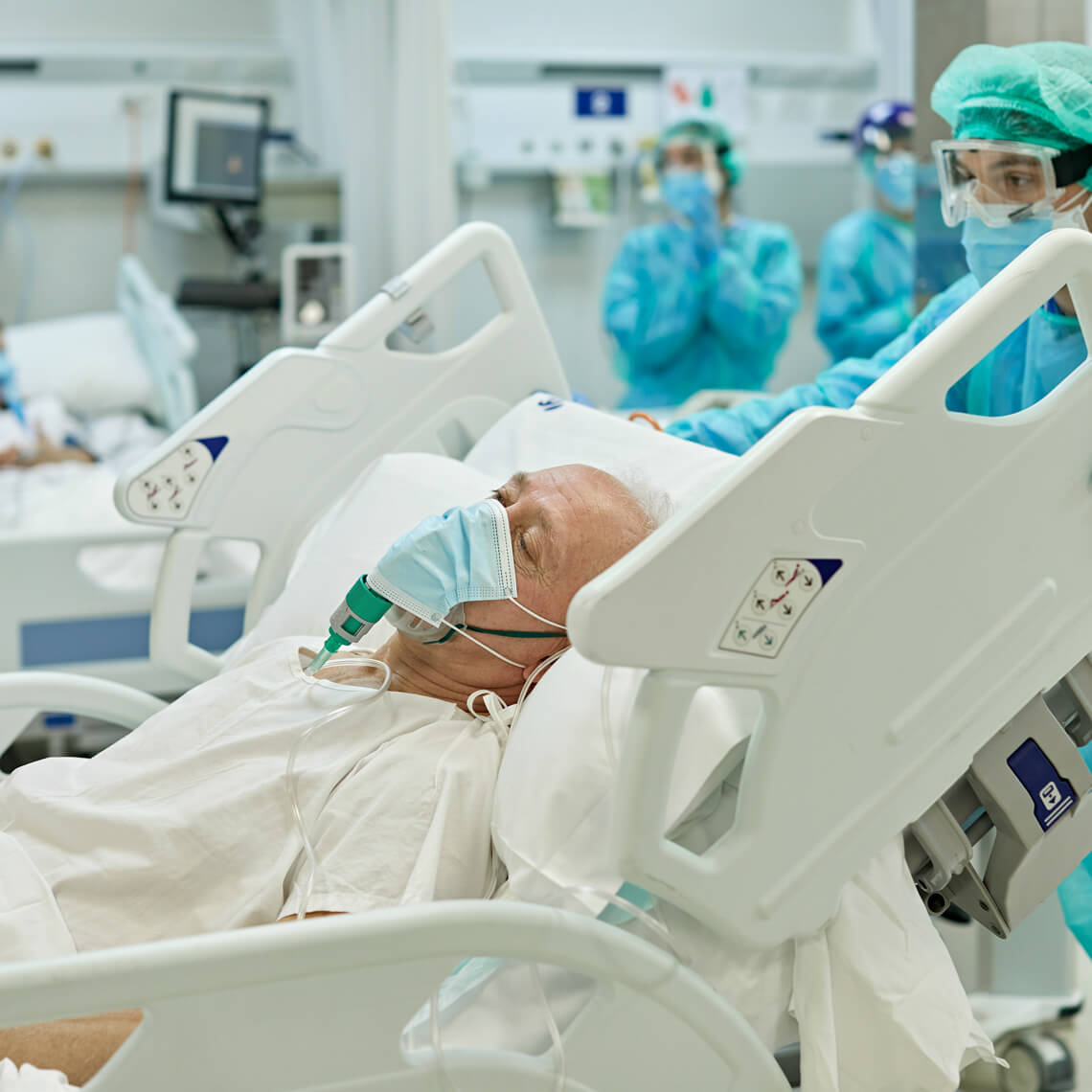 Senior Caucasian man on ventilator lying in hospital bed with eyes closed as medical professional wheels his bed past camera.