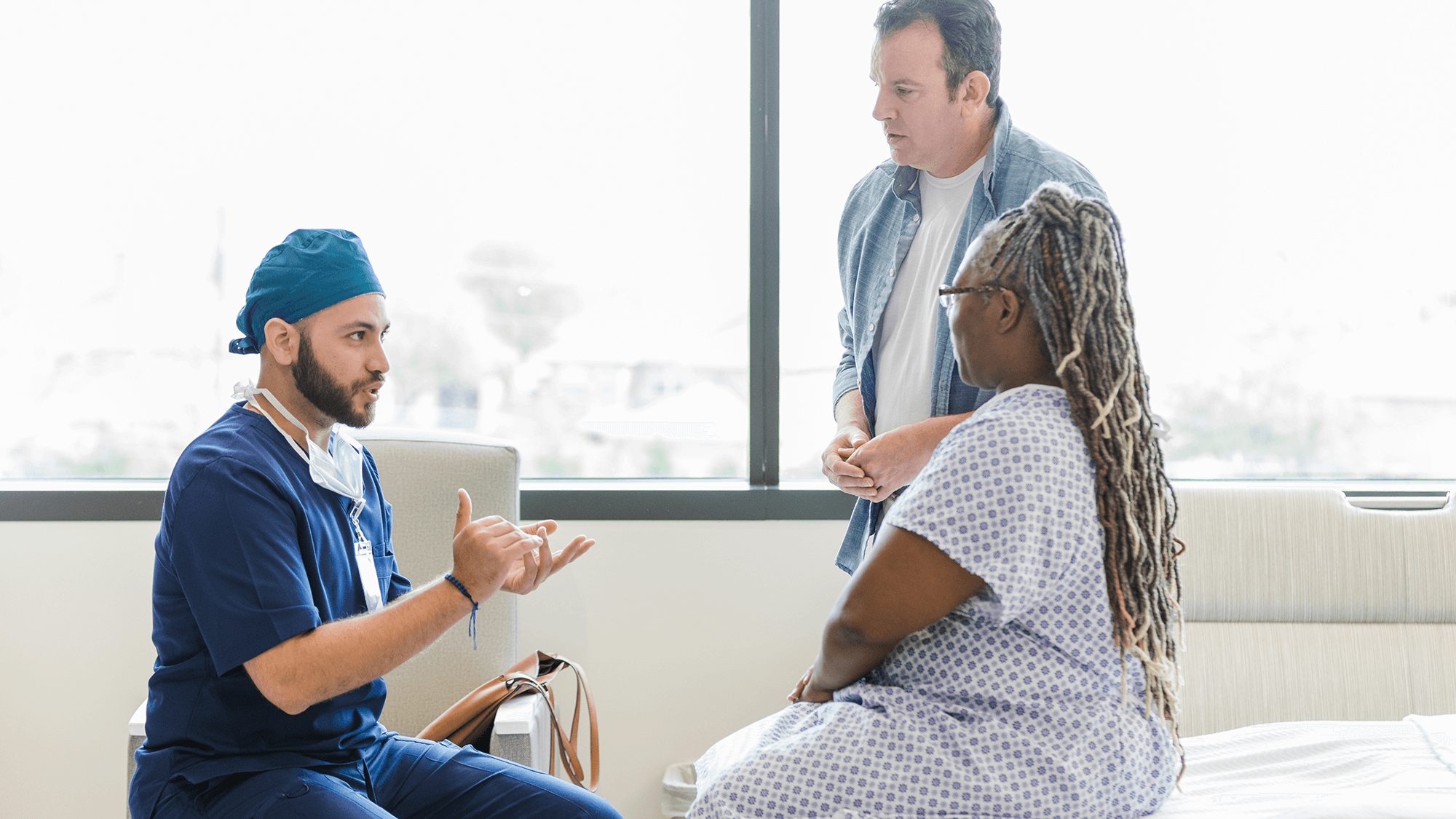 person in scrubs talks to a woman in a johnny who is seated on a hospital bed; a man stands beside her