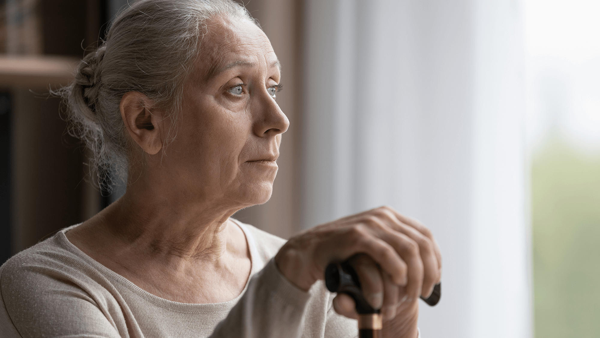 Elderly white woman in bun holds cane and looks thoughtfully out a window