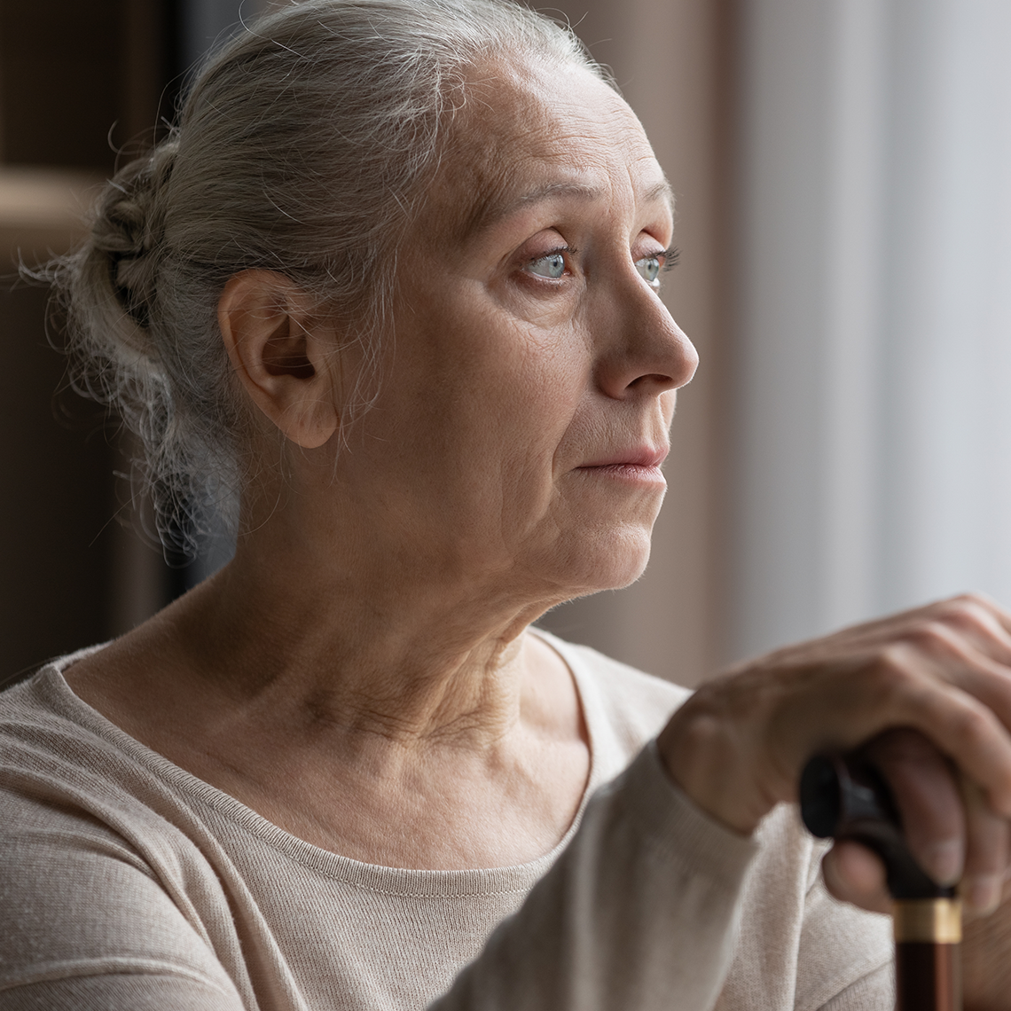 Elderly white woman in bun holds cane and looks thoughtfully out a window