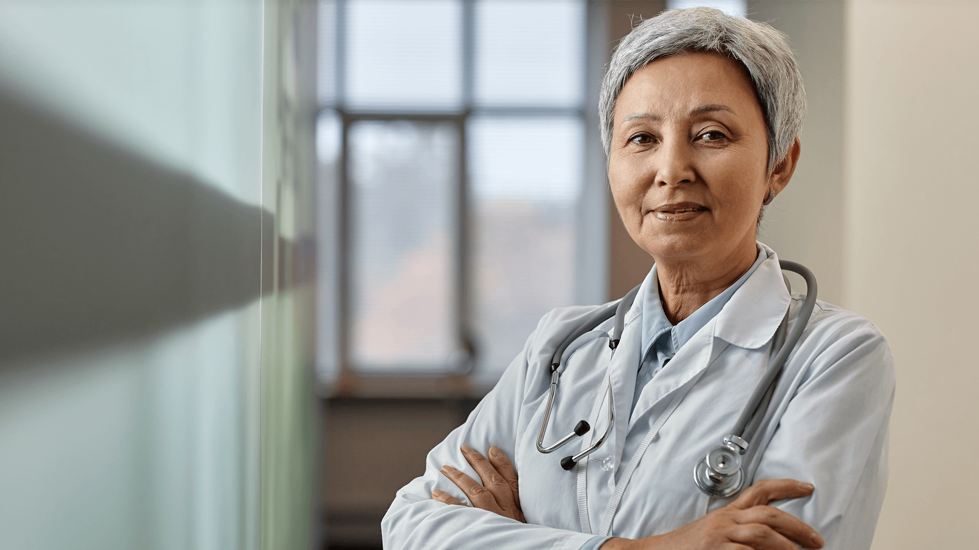 A woman in a doctor's coat stands by a window, looking confidently at the camera