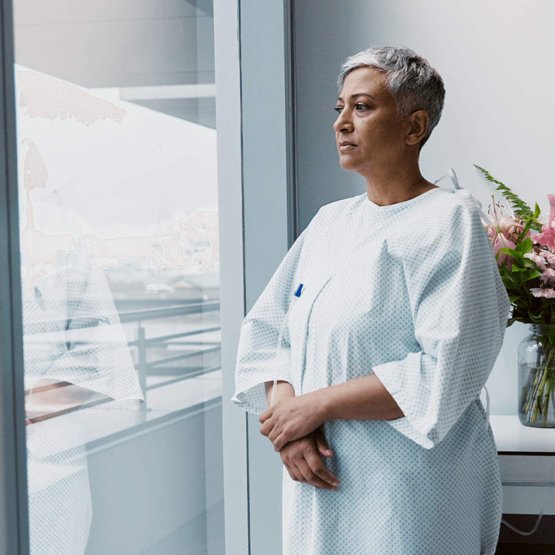 A patient in a hospital gown stands by a window, gazing outside. A colorful bouquet and a 