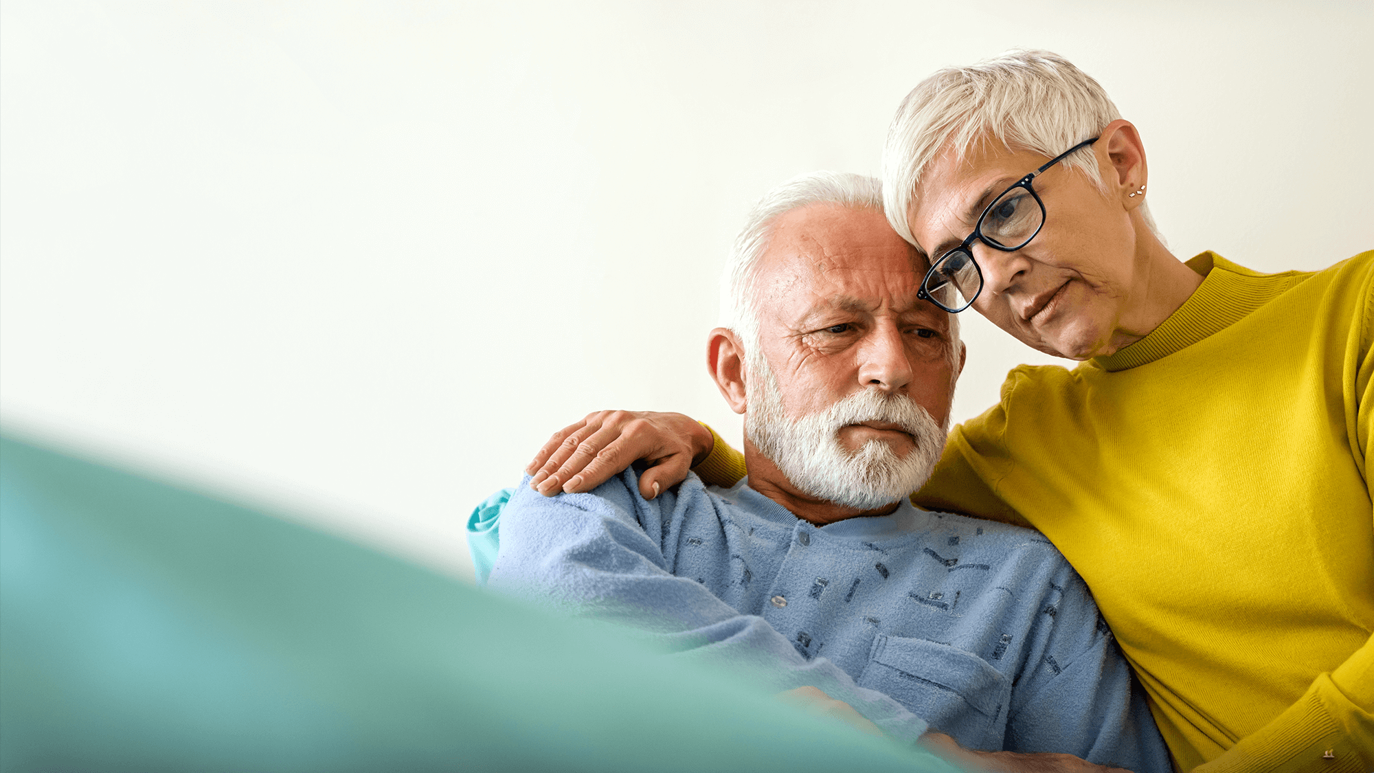 A woman gently rests her hand on the shoulder of an elderly person, both smiling, in a cozy setting with soft colors