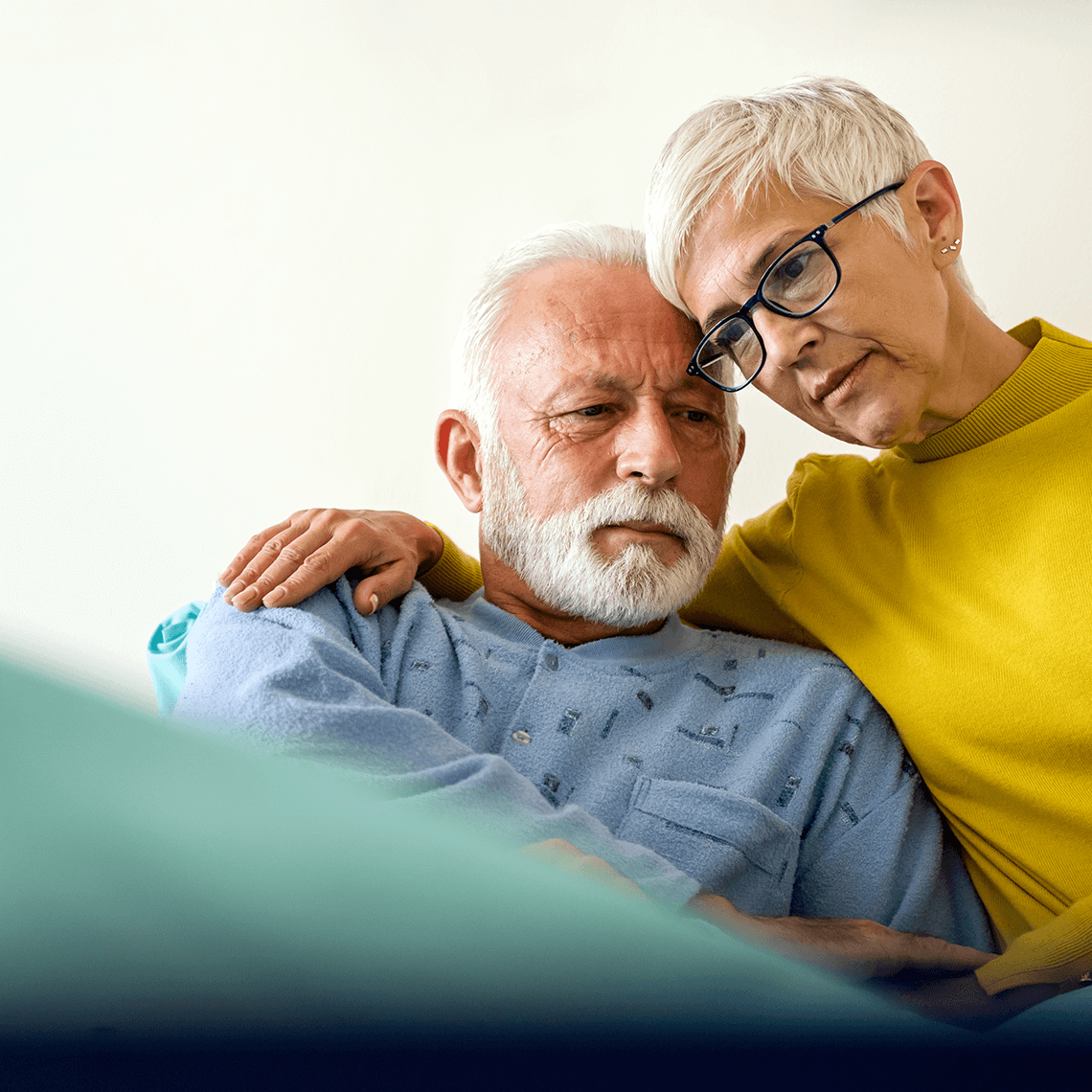 A woman gently rests her hand on the shoulder of an elderly person, both smiling, in a cozy setting with soft colors