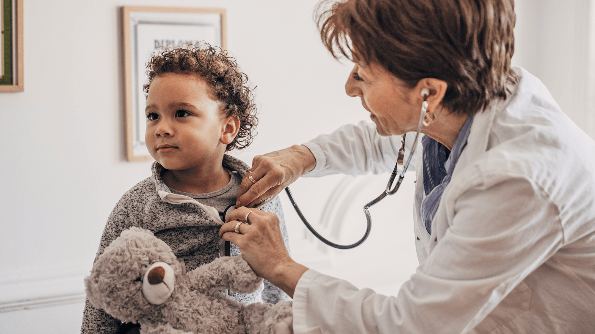 Little curly-haired boy with teddy bear gets heartbeat measured by female doctor