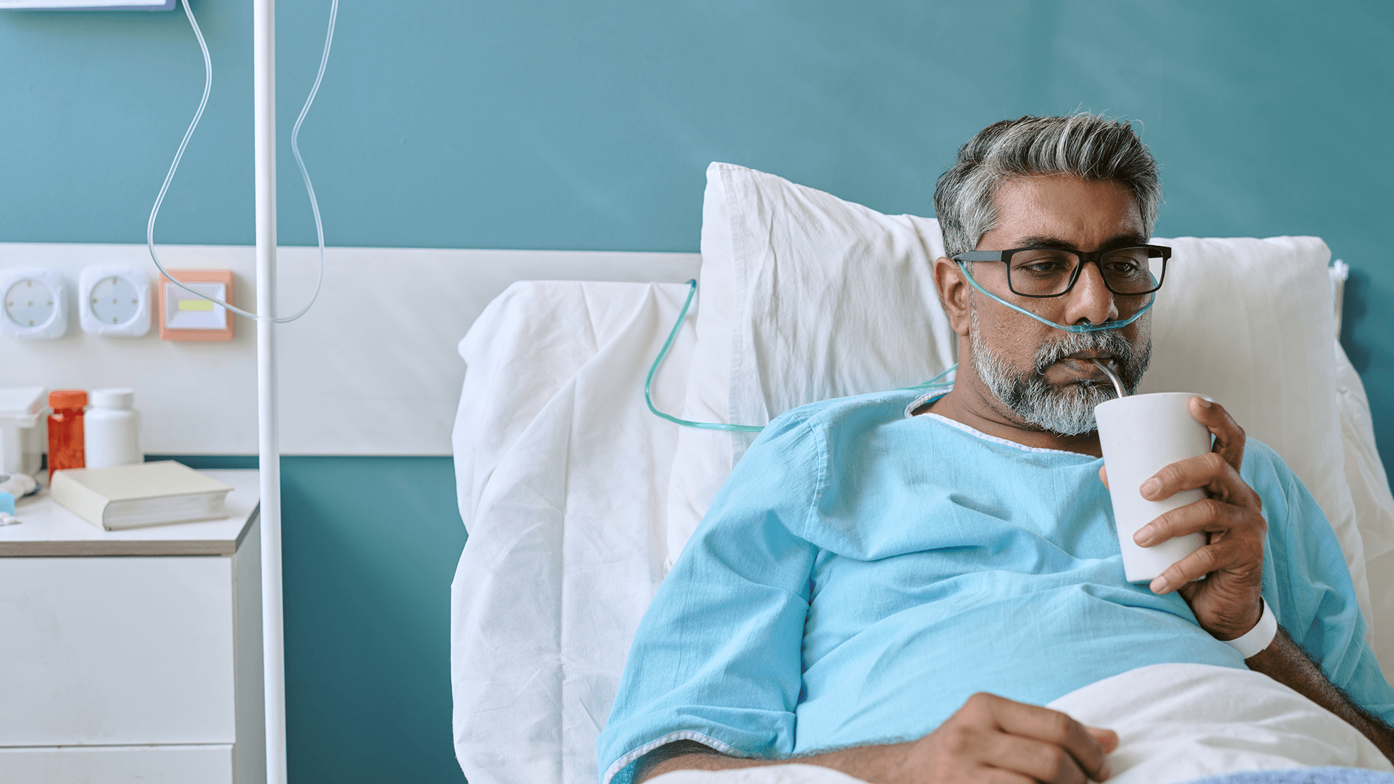 Bespectacled middle aged middle eastern male in hospital gown sipping a drink