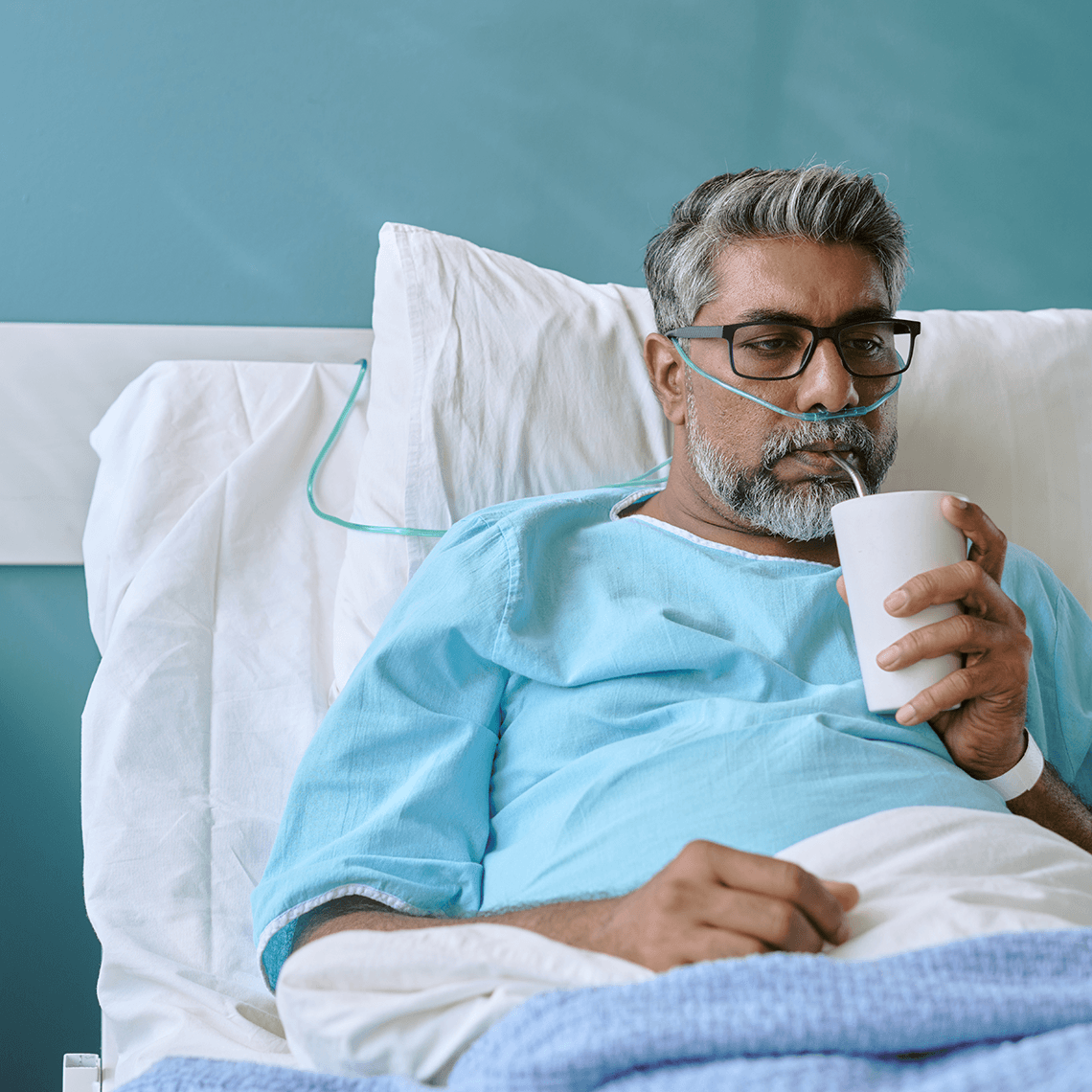 Bespectacled middle aged middle eastern male in hospital gown sipping a drink