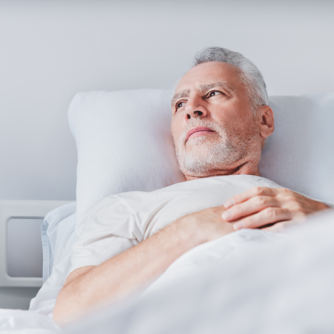 white bearded man staring wistfully off in distance while in bed against whitish grey background