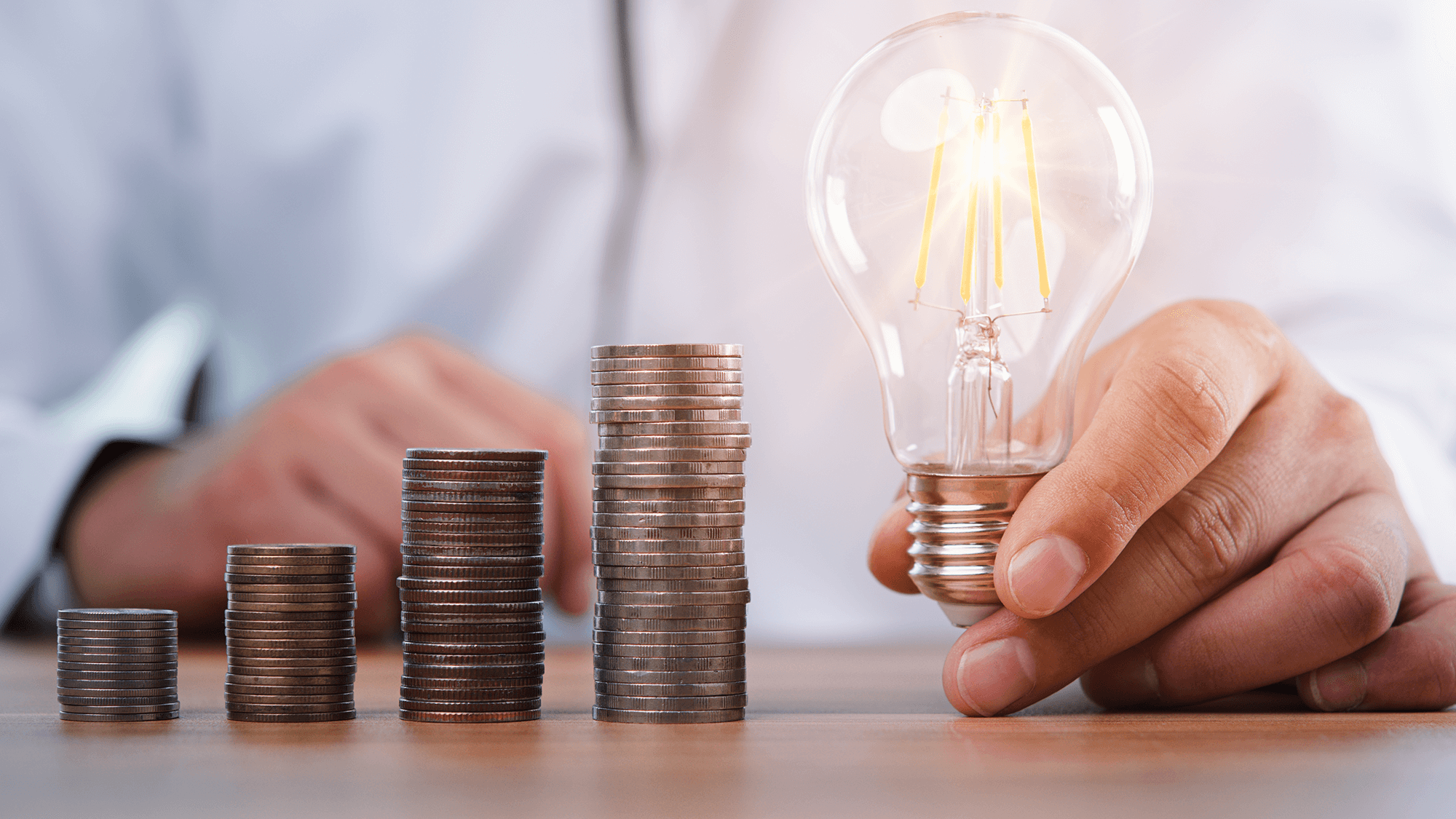 Close up of four increasing stacks of coins on a desk in front of a person wearing white. They are holding a lightbulb to the right of the tallest coin stack.