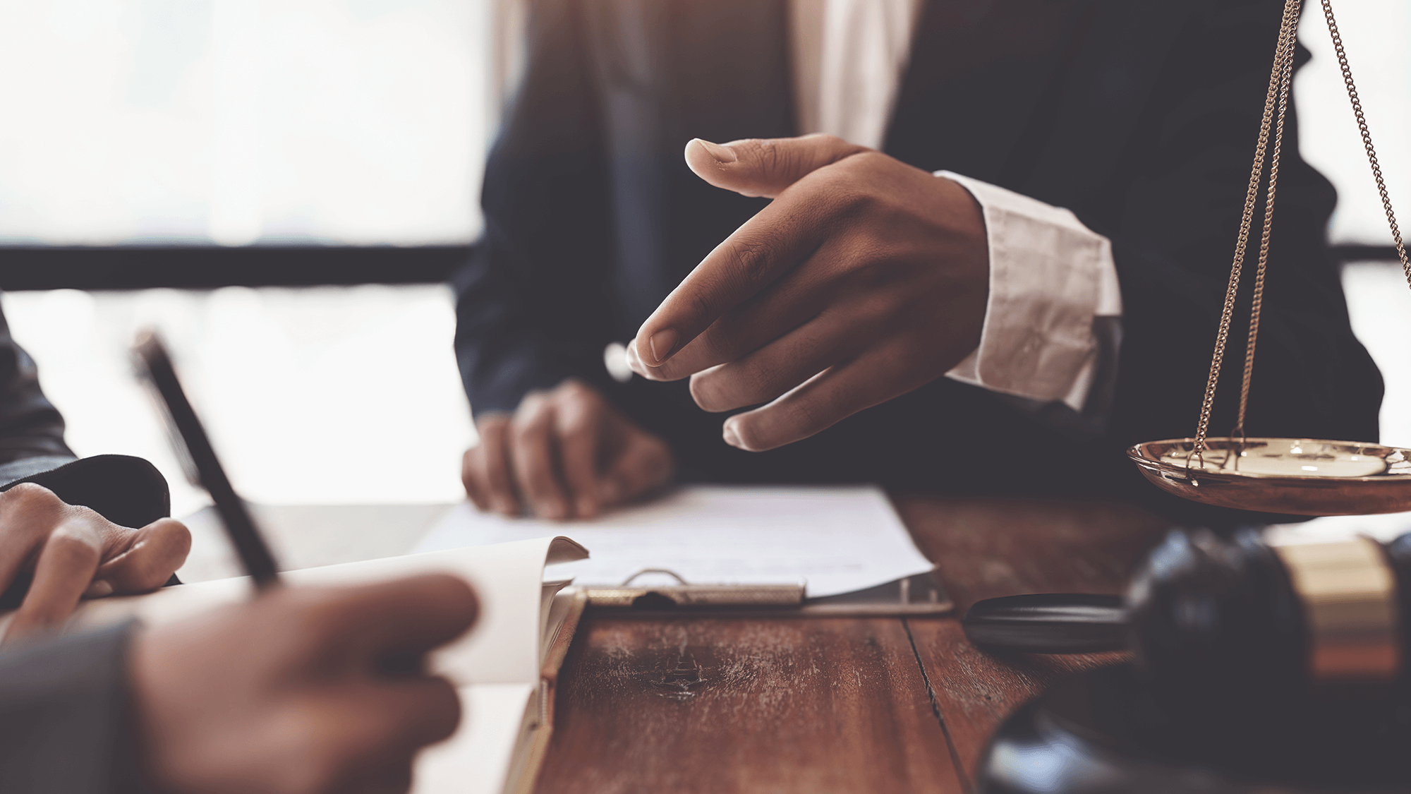 A close-up of a hand gesturing during a discussion, with legal documents and a gavel on a wooden table in a professional setting.