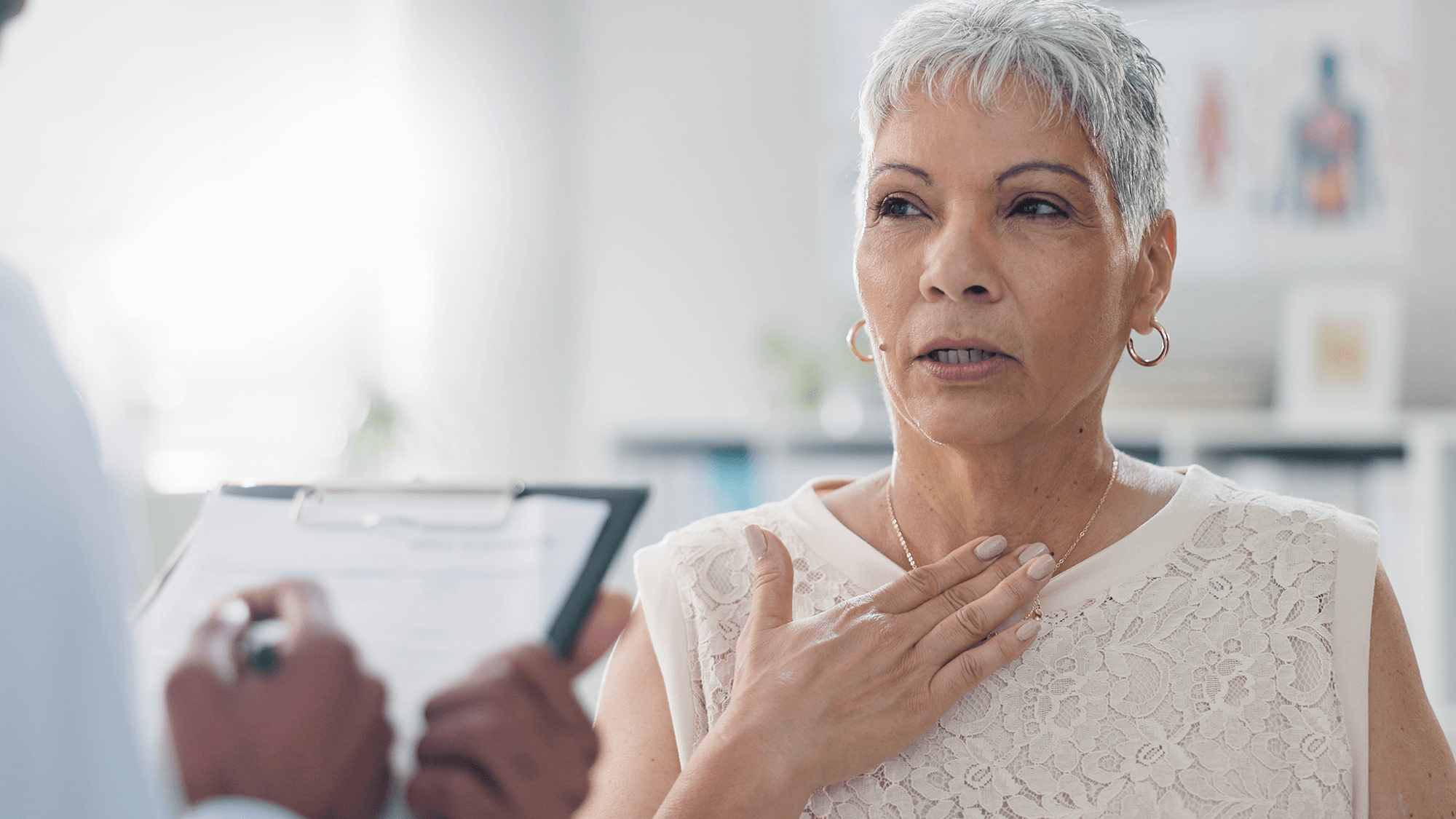 A woman with short gray hair is holding her hand to her chest while listening to a healthcare professional. A clipboard is held in the foreground.