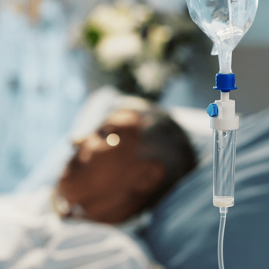 A close-up of an intravenous (IV) drip with a blurred patient lying in a hospital bed in the background